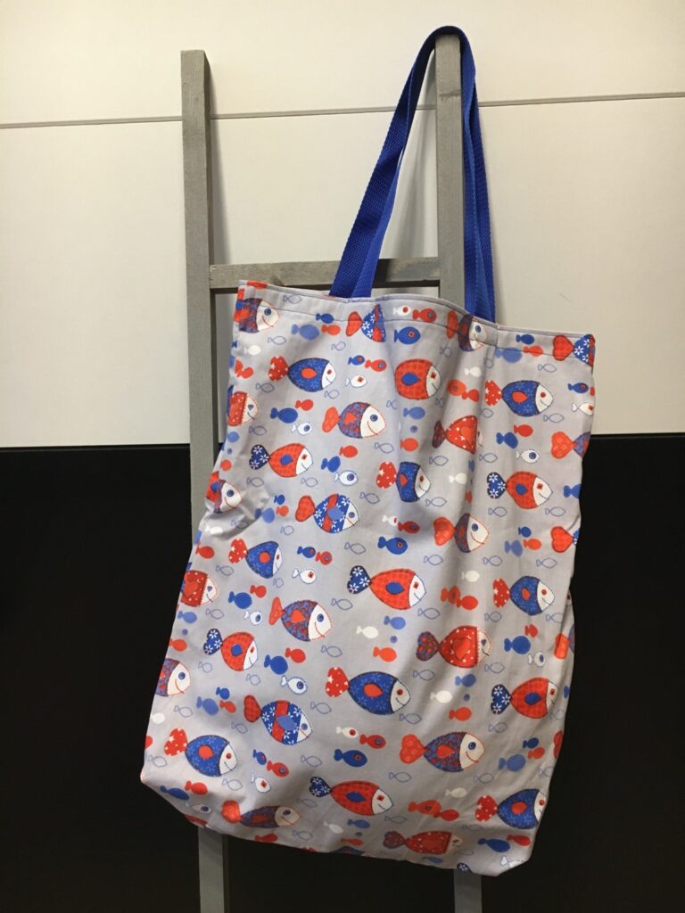 Stofftasche mit buntem Fisch-Print und blauen Henkeln an einer Leiter