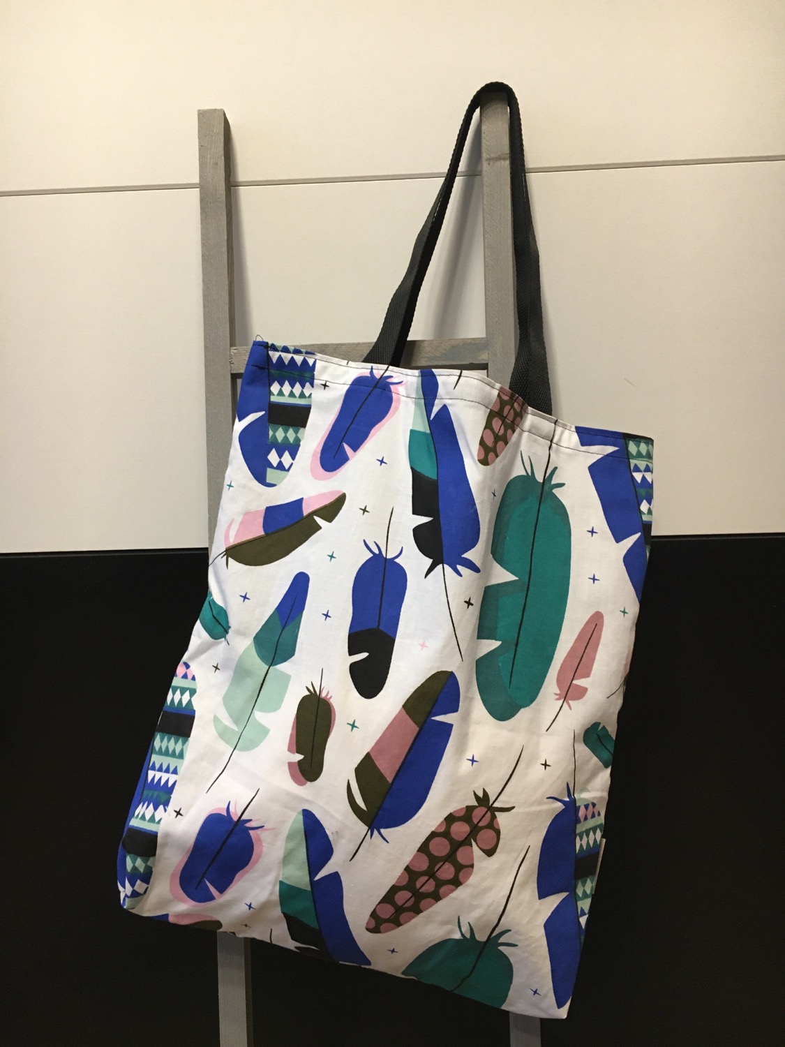 Stofftasche mit buntem Feder-Print und dunklen Henkeln an einer Leiter